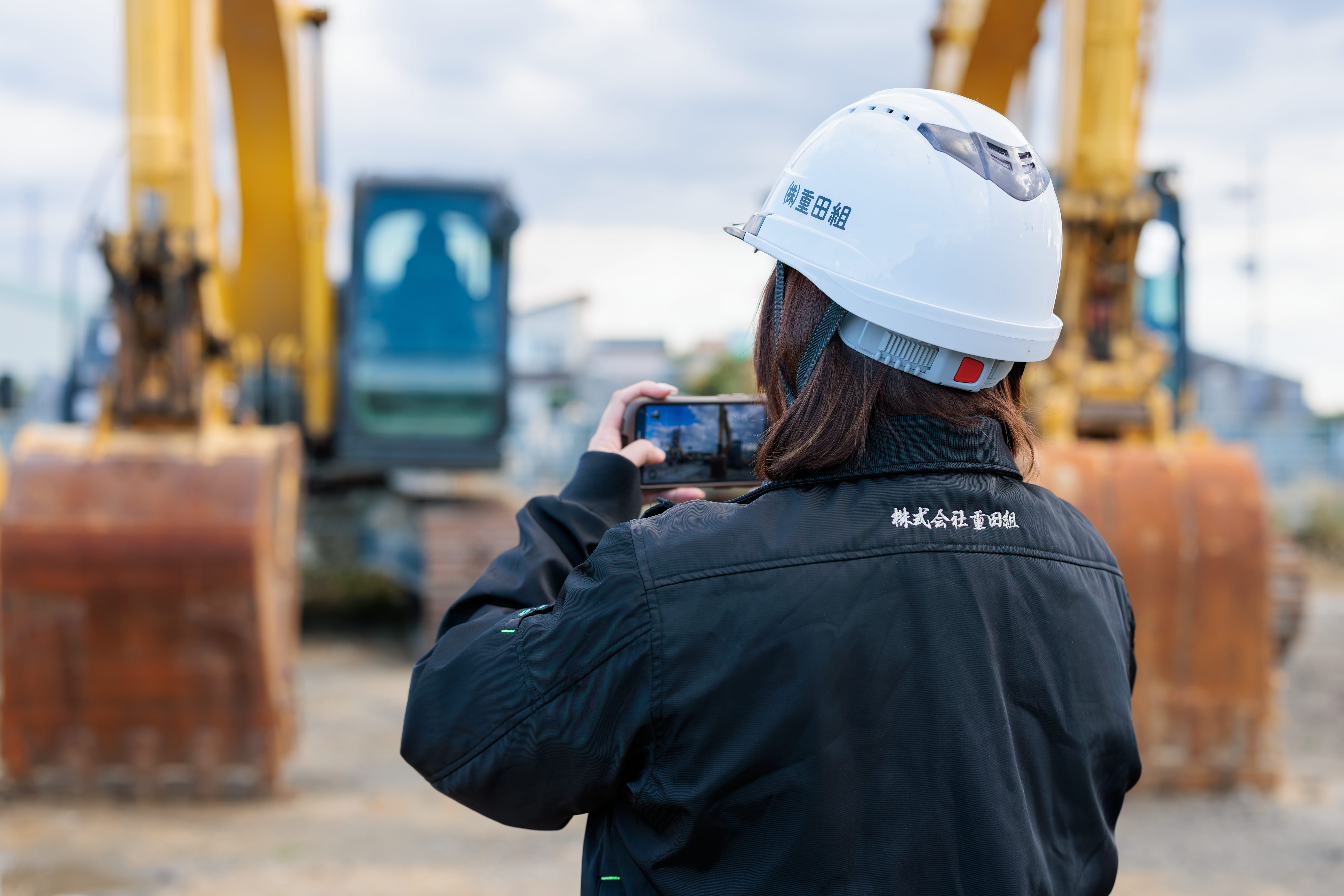 建設現場でヘルメットを着用し、スマートフォンで写真を撮る作業員の後ろ姿