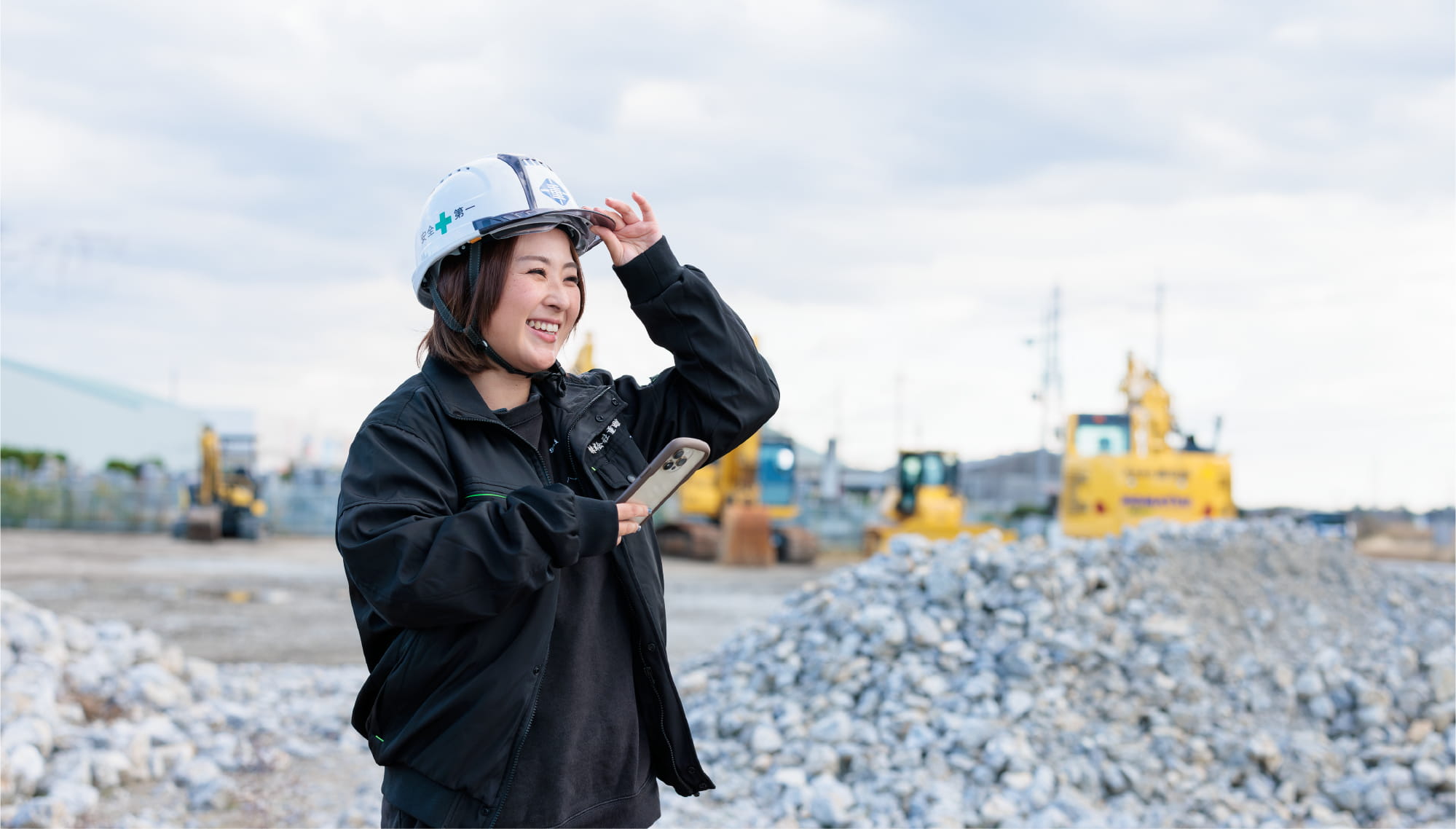 ヘルメットをかぶった女性が建設現場で笑顔でスマホを持っている様子