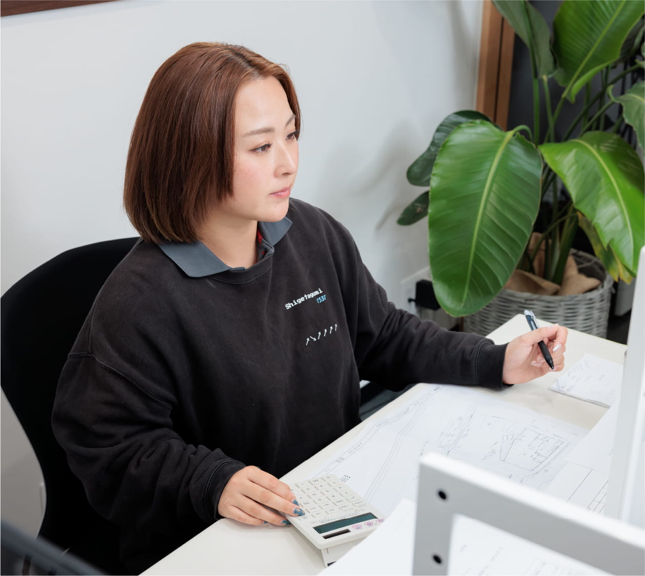 女性がオフィスで設計図を見ながら電卓を使い、計算や作業をしている様子