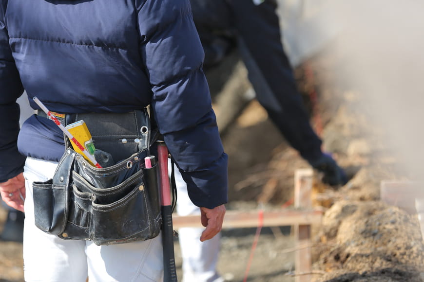 工事現場で作業する職人の腰に、多くの工具が入った黒い腰袋が掛けられている