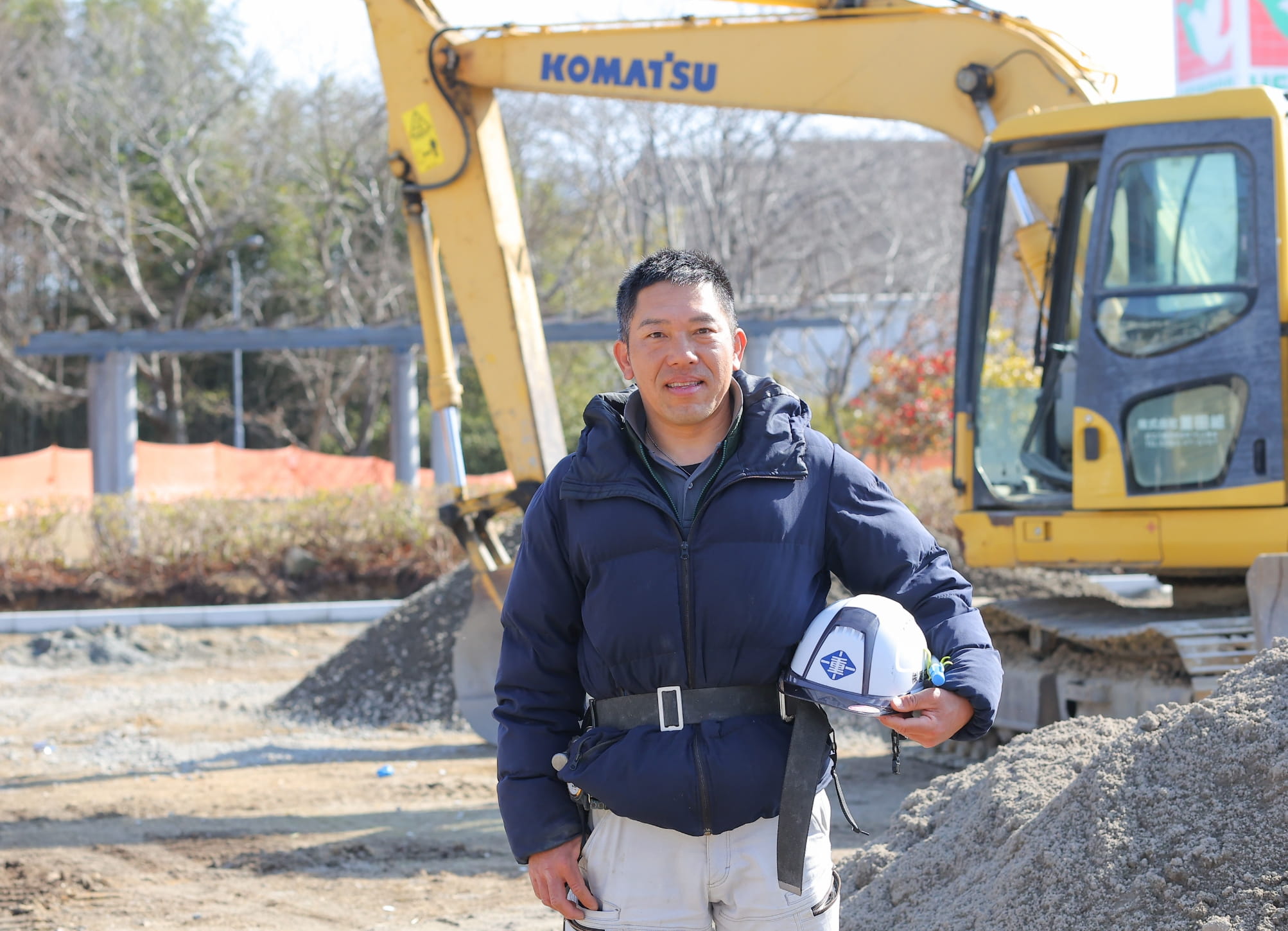 建設現場で働く男性が、重機を背景にヘルメットを持ちながら微笑んでいる