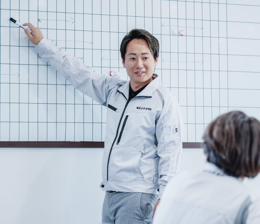 ホワイトボードの前で作業着姿の男性が指し示しながら説明をしている場面