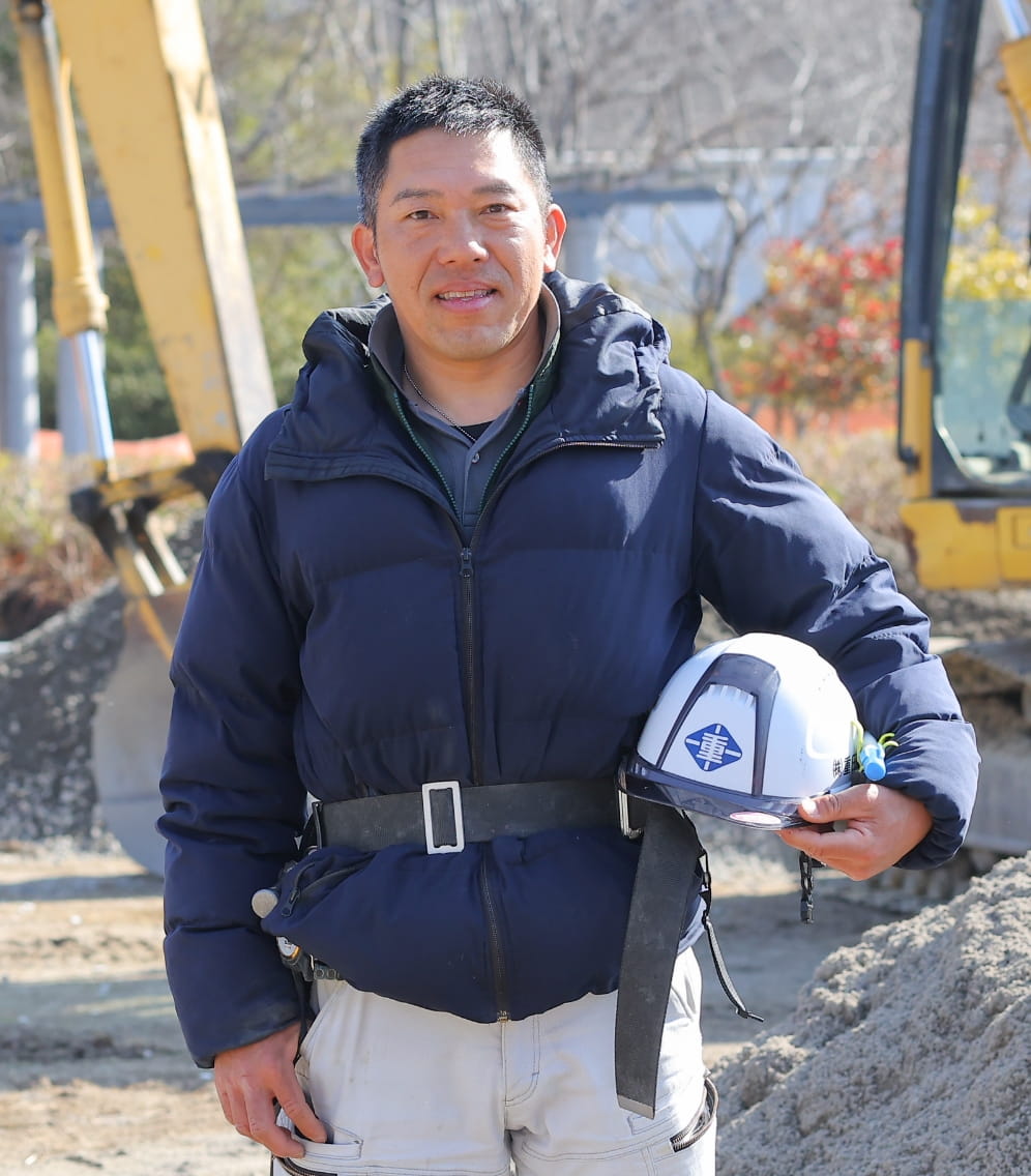 建設現場で働く男性が、重機を背景にヘルメットを持ちながら微笑んでいる